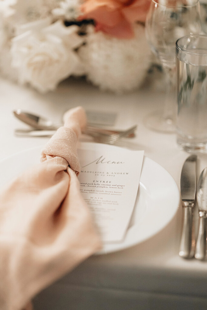 Details of the table setting show white plates, pink knotted napkins, silver cutlery, florals and menus