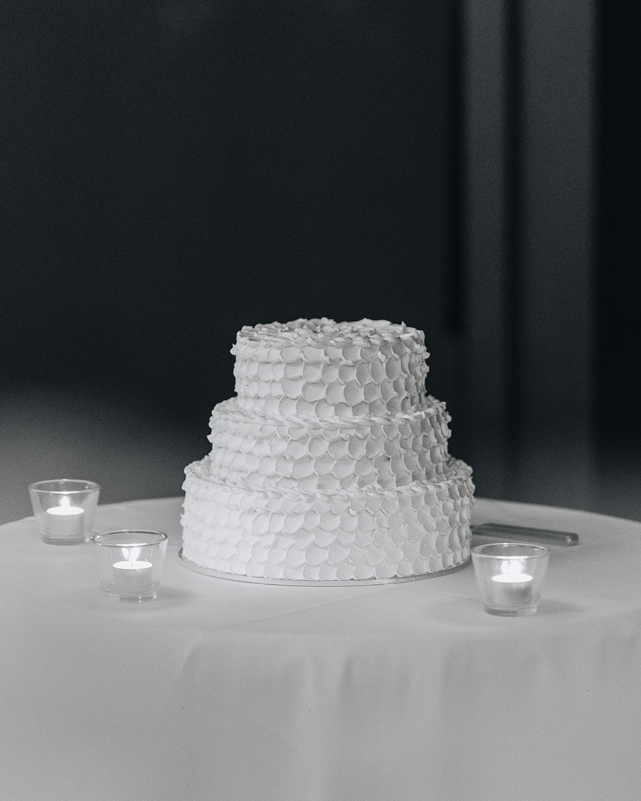 The wedding cake is an elegant 3 tiered cake, sitting on a round table with tealight candles
