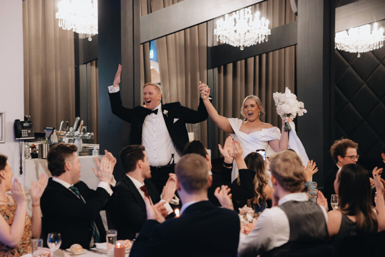 The bride and groom enter the reception, holding hands and cheering as guests are seated before them, clapping.