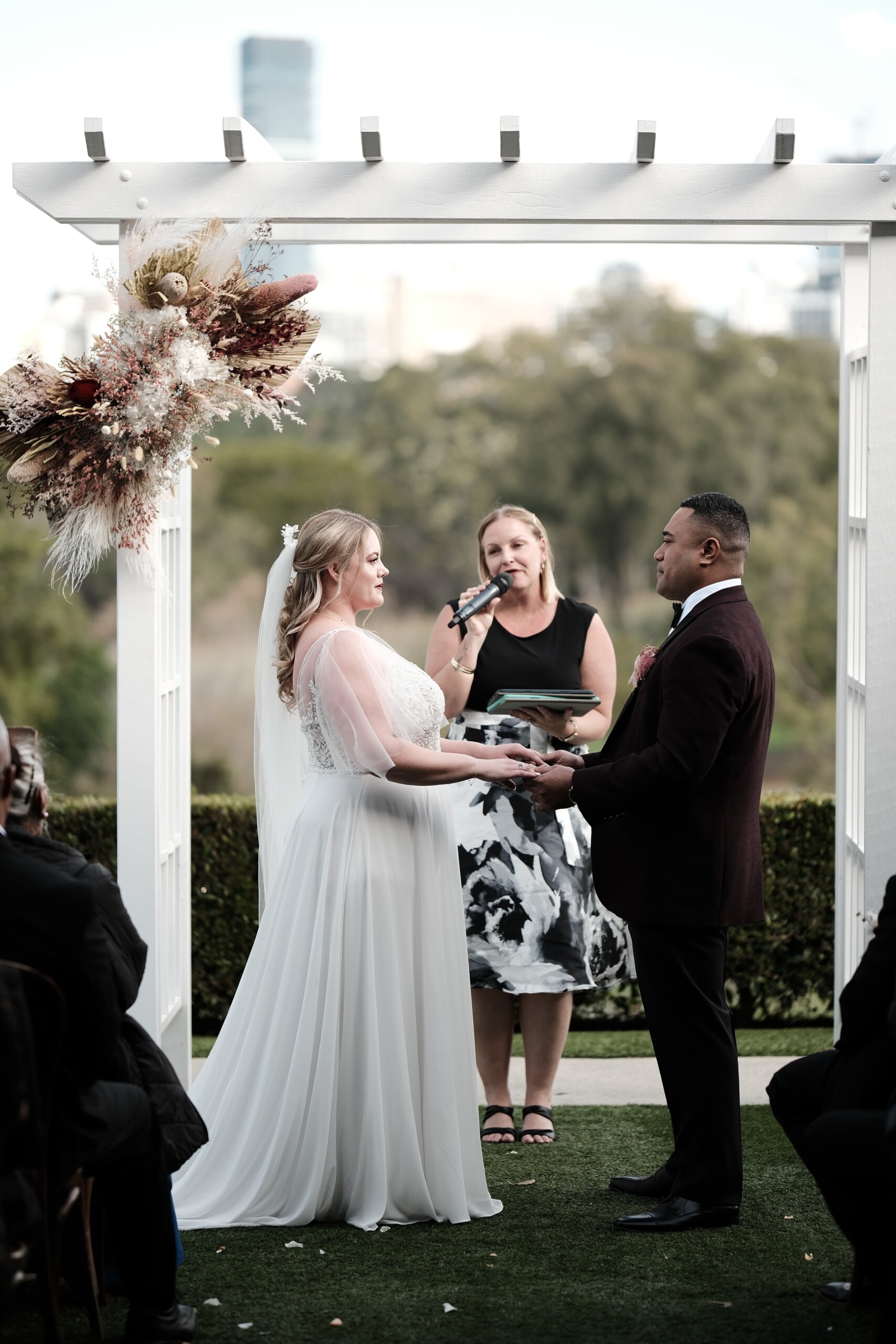 Rose and Taape stand together holding hands at the alter, as the celebrant speaks into a microphone. 