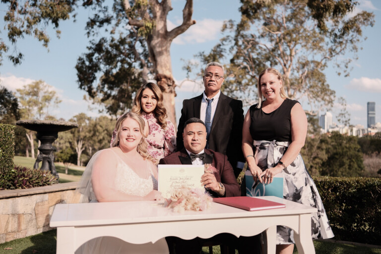 Rose and Taape sit at a table, holding their marriage certificate, surrounded by witnesses.