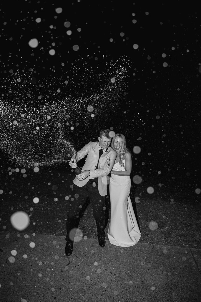 Bride and groom pop a champagne bottle towards the camera, at night time.