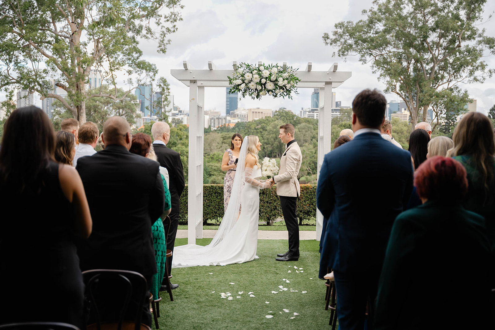 Bride and groom stand at the alter, underneath a white arbour and white florals. Guests are standing nearby and the celebrant holds a microphone. The parklands city skyline is in the background.
