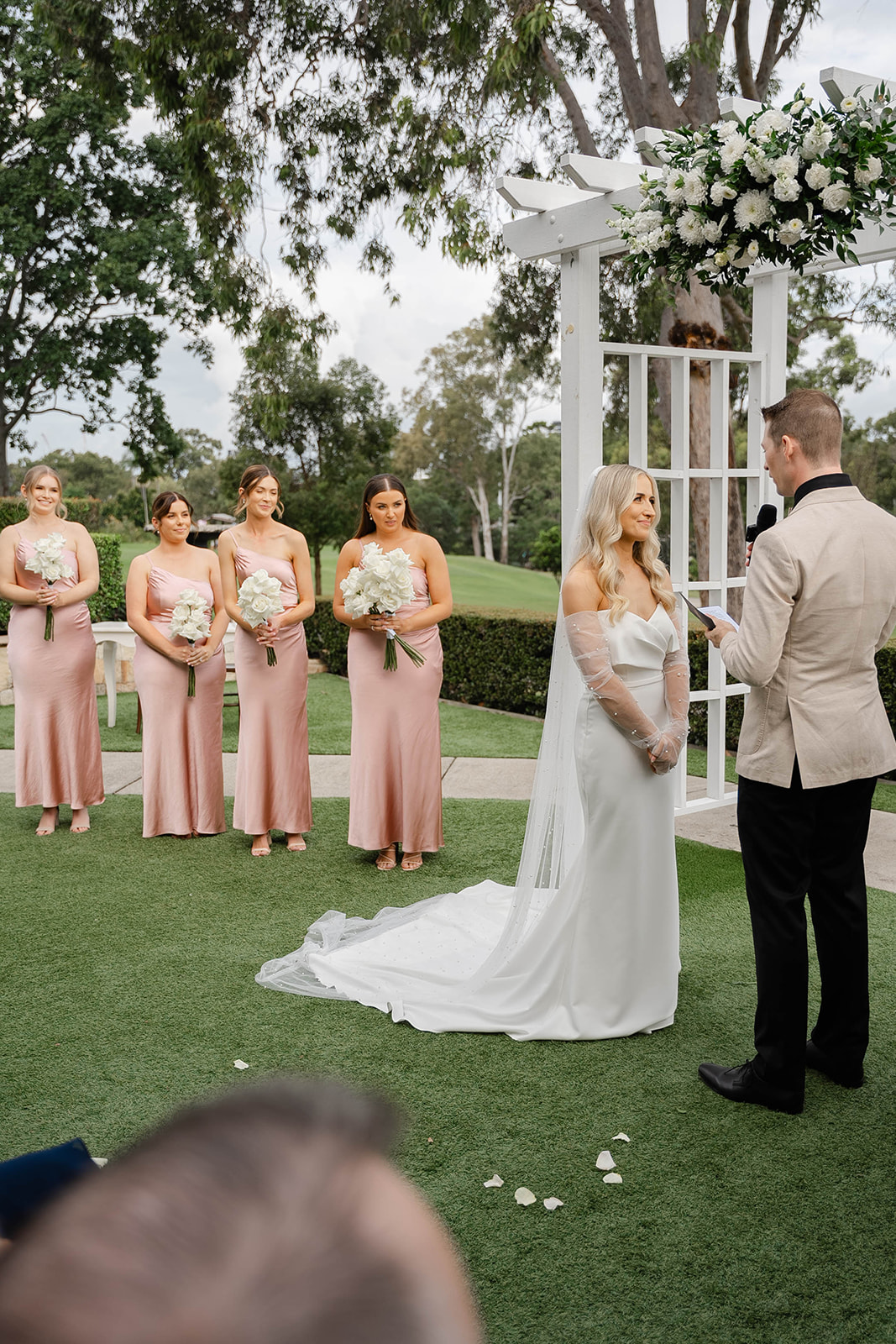 Bride and groom stand at the alter, while the groom reads his vows. The four bridesmaids stand near the bride wearing blush pink dresses, holding white bouquets.