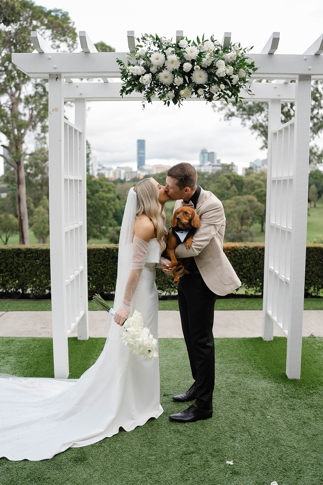 The bride and groom kiss at the alter, with their Cavoodle dog in the grooms' arms. The bride holds her bouquet of flowers.
