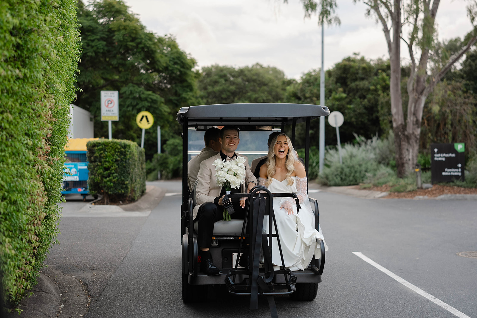 The bride and groom ride in the back of a golf cart, laughing and smiling, as they drive to get photos taken.