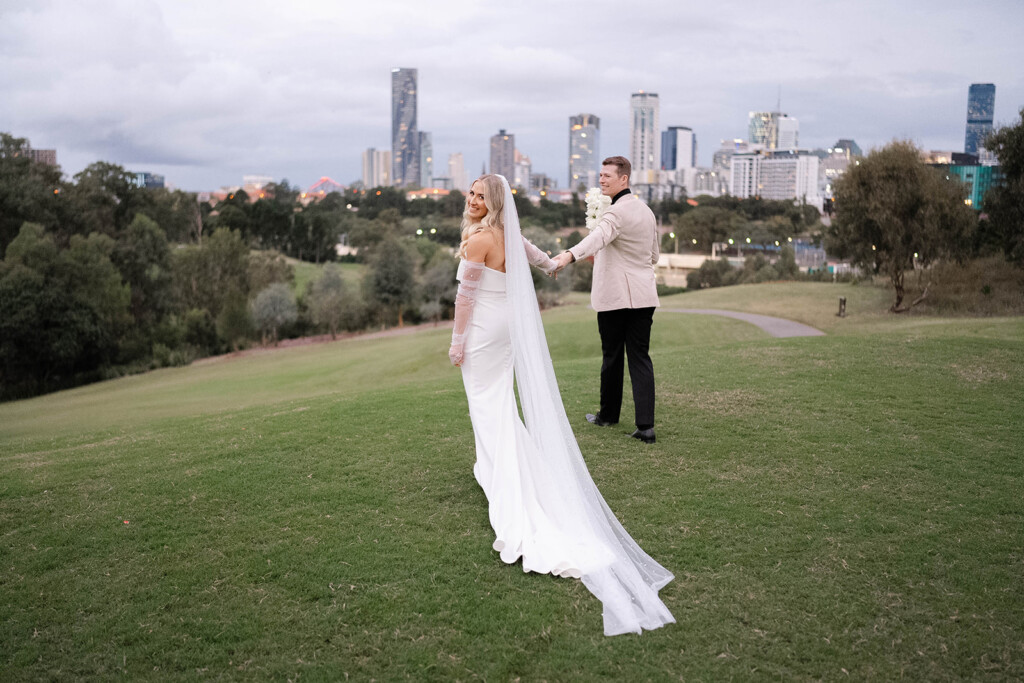 Bride and groom walking together in the parklands, with the city skyline on the horizon. The bride is looking back at the camera with her veil flowing behind.