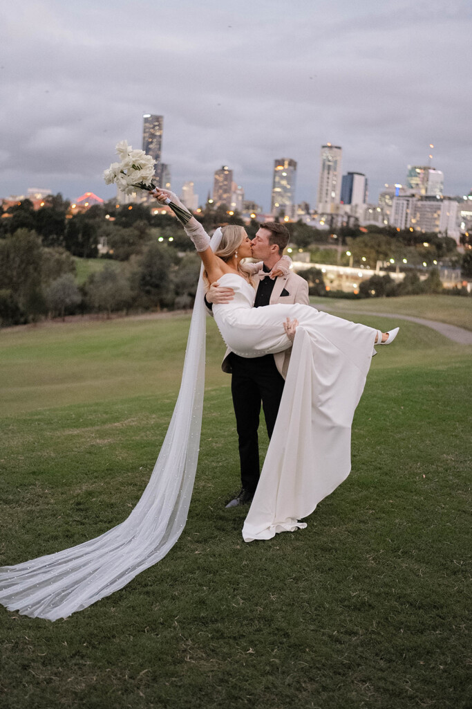 The groom holds bride in his arms, upon dusk with the city sparkling behind. The Bride holds her boutique in her hand, raised above her head. They are sharing a kiss.