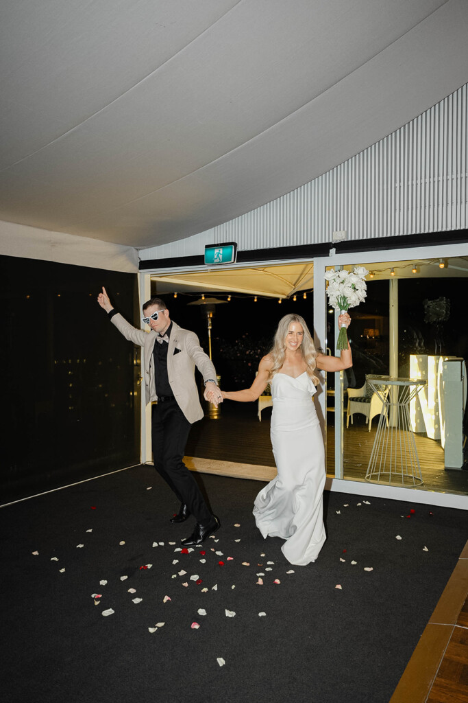 Bride and groom walk into the reception holding hands, there are flower petals on the ground.