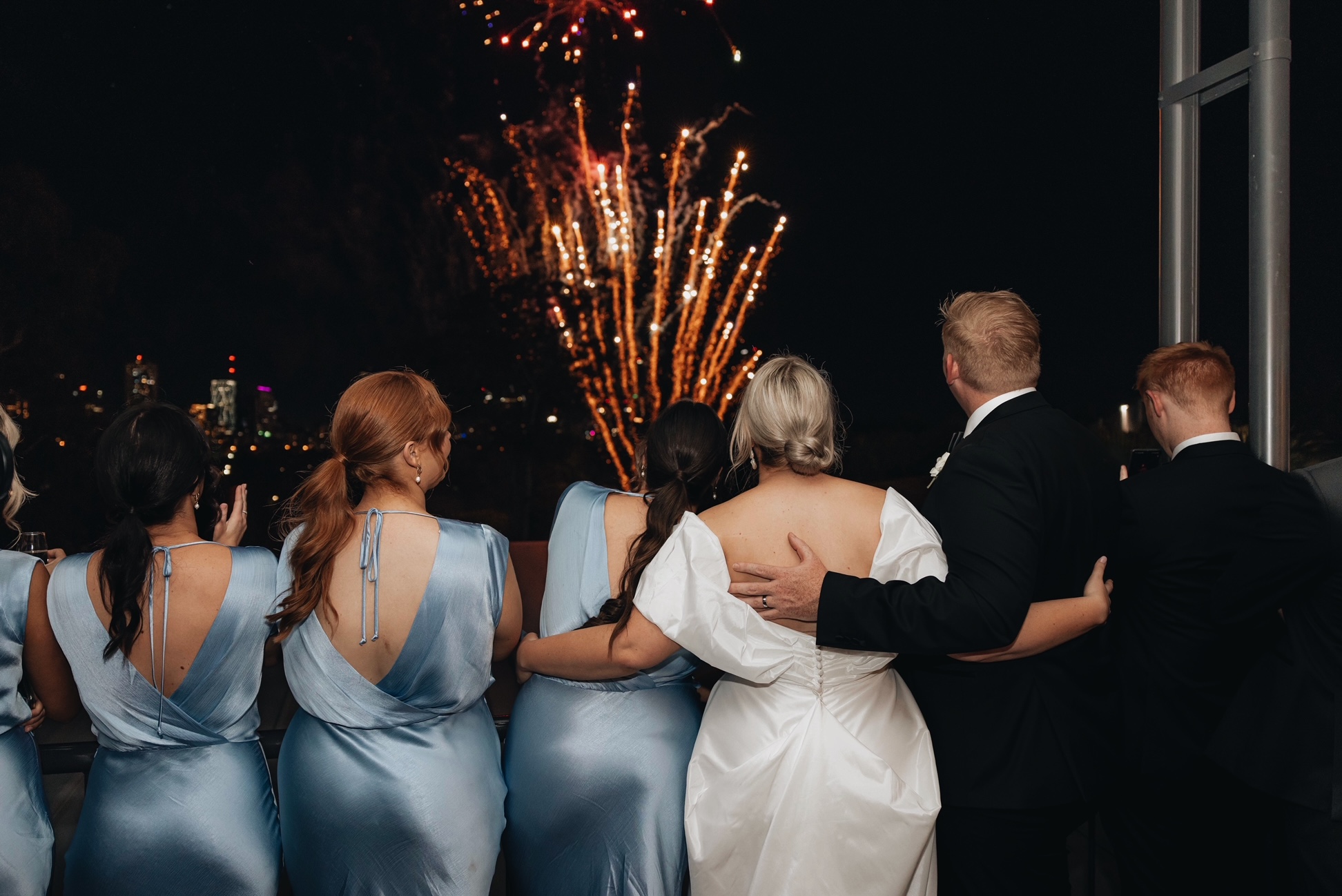 The bridesmaids and wedding couple stand on the balcony, watching fireworks above
