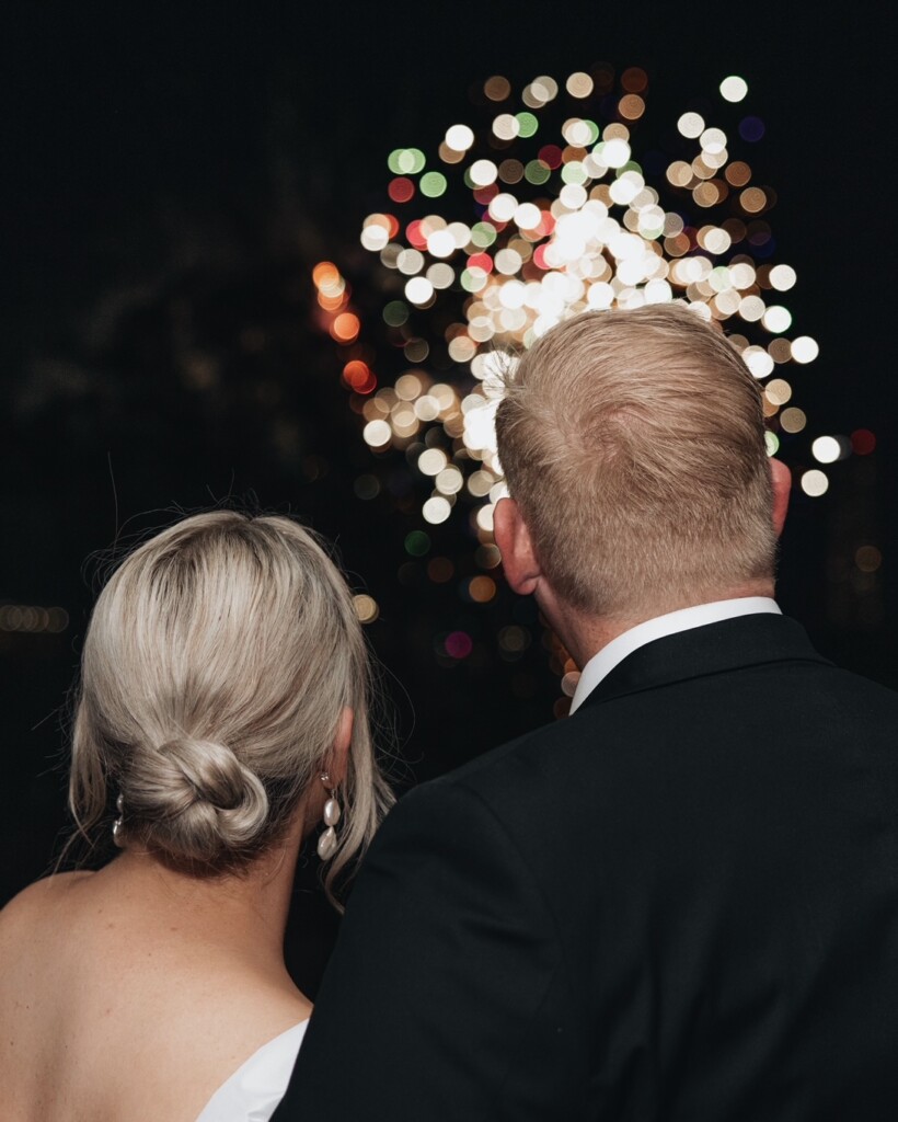 The wedding couple stand together as they watch fireworks in the night sky.