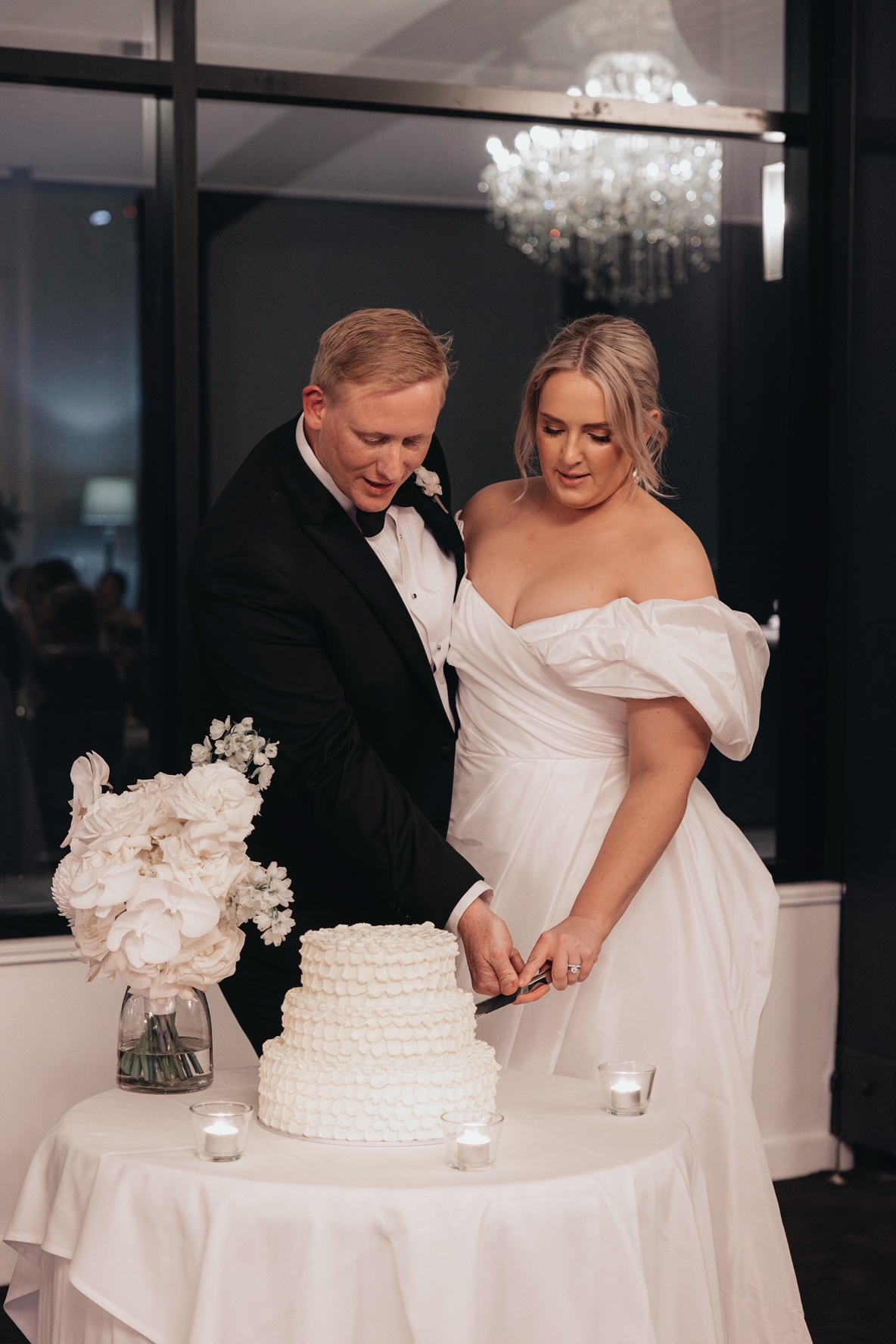 The bride and groom cut the cake together. A floral arrangement sits beside the cake.